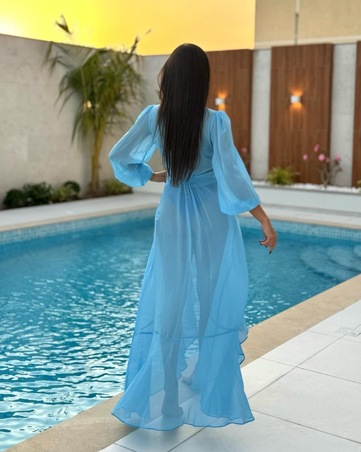 Woman in a blue dress standing by a pool at sunset.